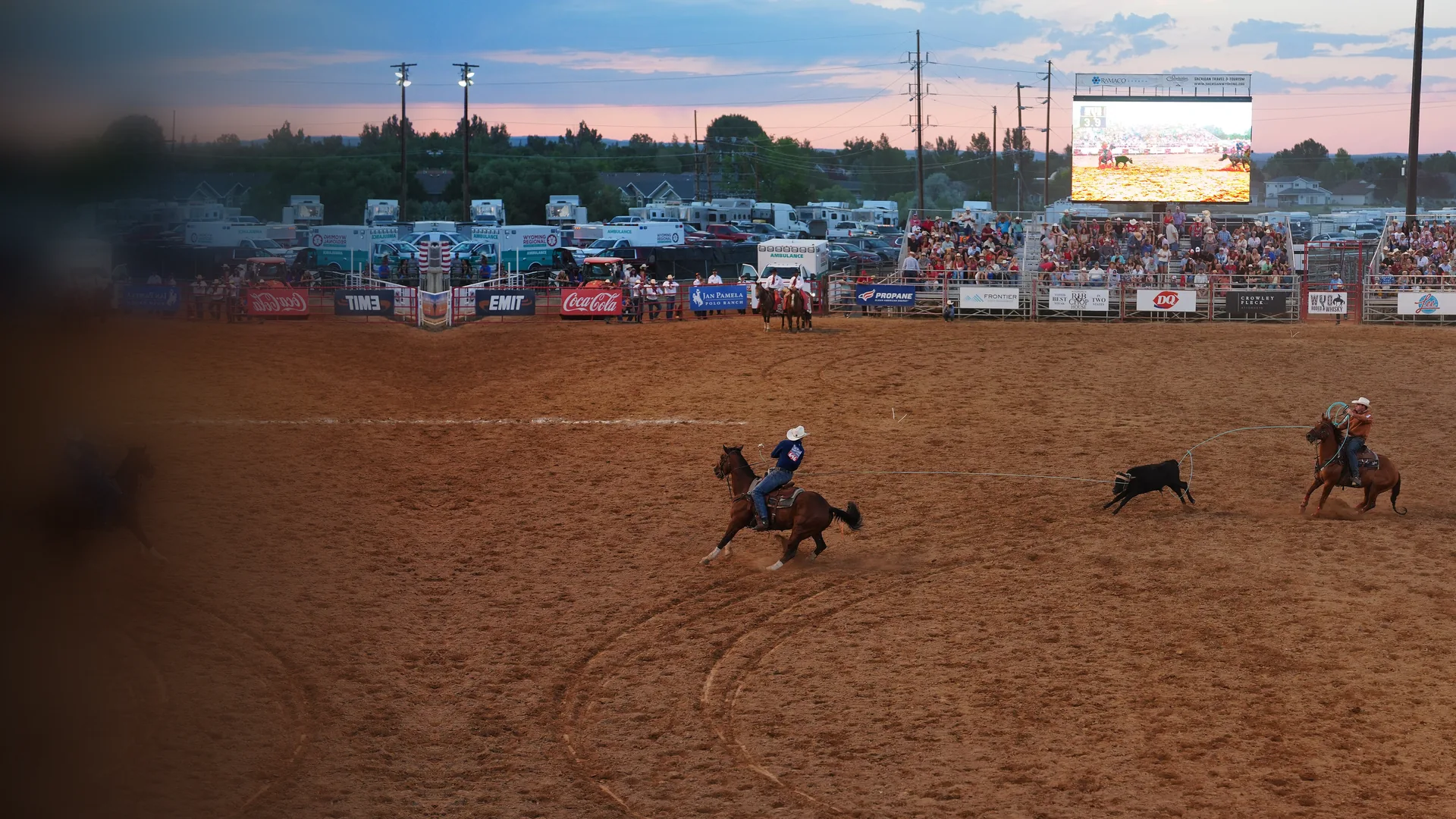 Zwei Reiter zu Pferd, die in der Abenddämmerung einen laufenden Steer über eine staubige Rodeo-Arena rope, mit Publikum auf den Tribünen und einer großen Leinwand über dem Zaun.